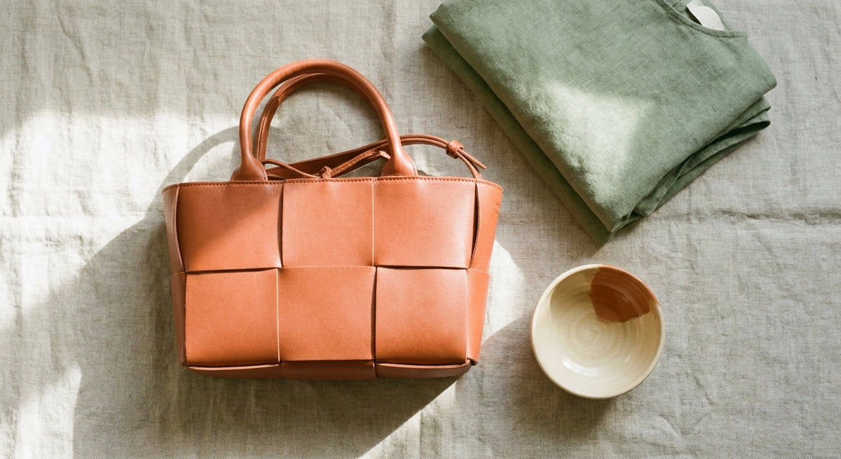 A sunlit flat lay featuring a woven leather handbag, folded natural fabric garment, and artisan ceramic bowl arranged on a linen surface in warm earthy tones.